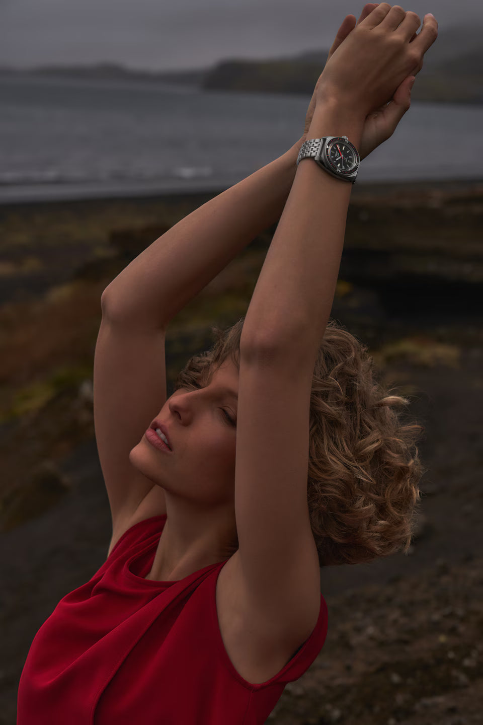 Person with outstretched arms by a body of water, wearing a red top and a watch.