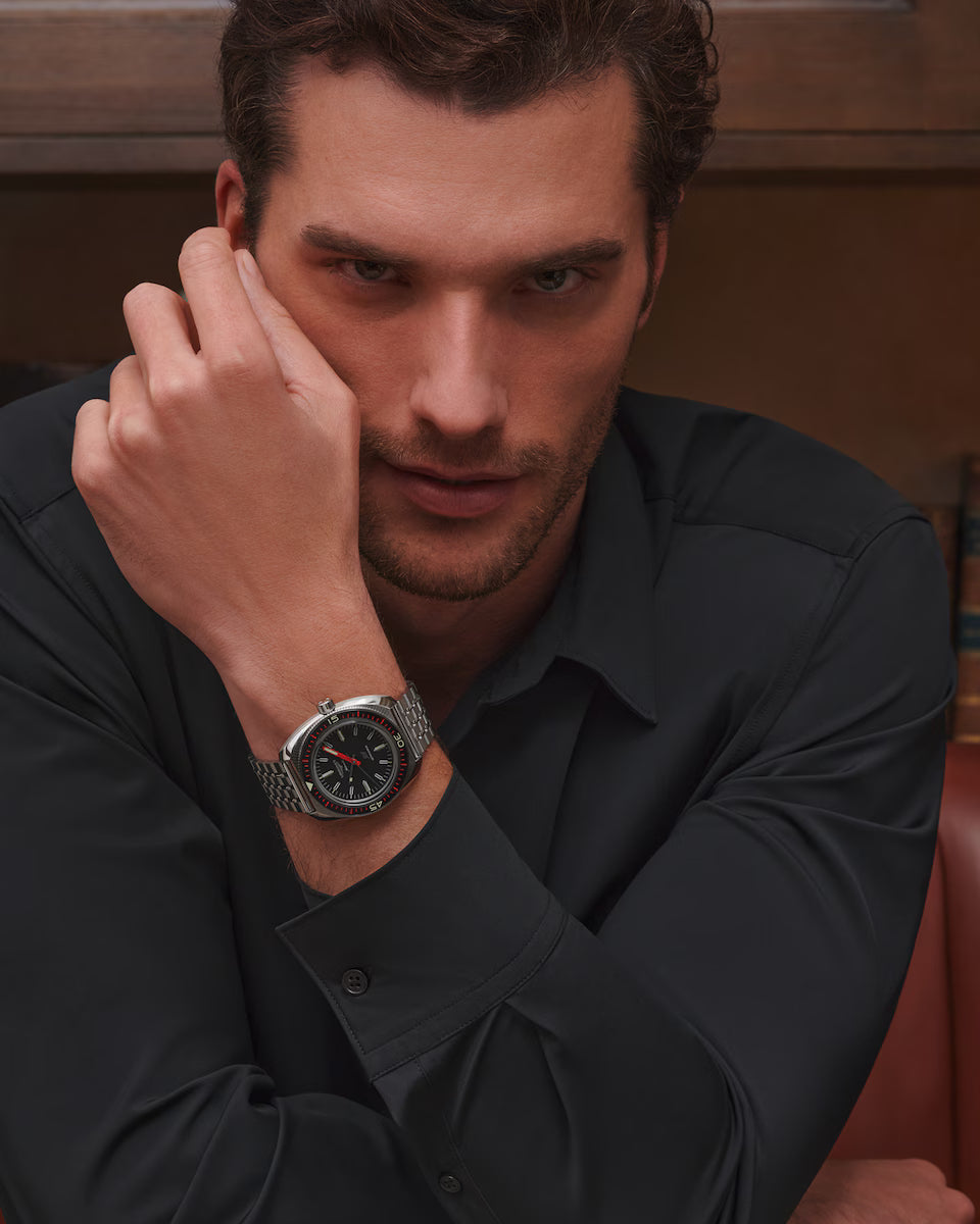 Man wearing a black shirt with a watch on his wrist, sitting in a chair.