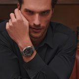 Man wearing a black shirt with a watch on his wrist, sitting in a chair.